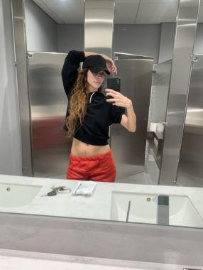 A woman wearing a black baseball cap, black sweatshirt, and red pants takes a selfie in the mirror of a restroom. Her right arm is resting atop her head and several scars are visible on her lower abdomen.